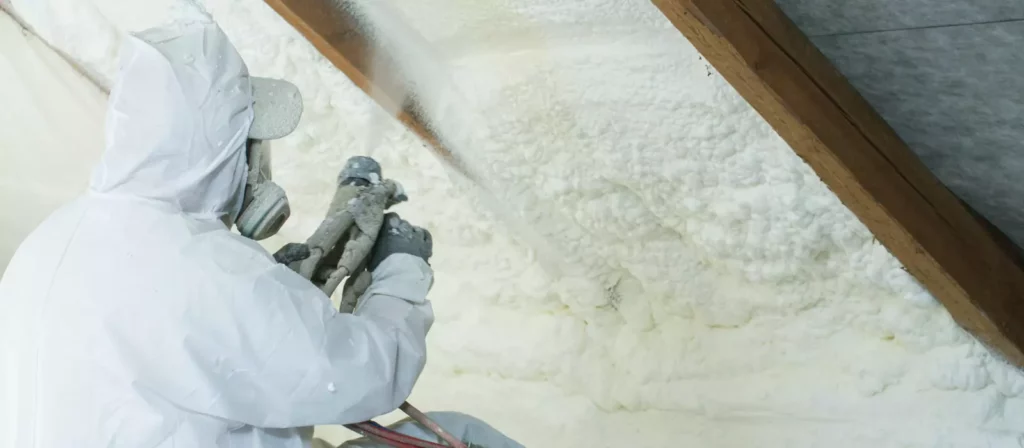 Spray foam installer in full hazmat suit spraying insulation into pitched roof rafters.